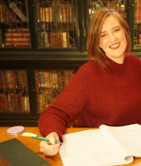 A woman with shoulder-length brown hair smiles at the camera. She is holding a pen over an open white notebook, with bookshelves behind her.