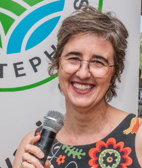 A woman with short gray-brown hair and glasses, holding a microphone and smiling.