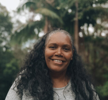 A dark-skinned woman with long curly black hair smiles at the camera, surrounded by trees