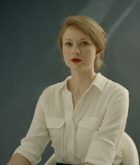 A strawberry-blonde woman with red lipstick and a white shirt sits up and looks directly at the camera
