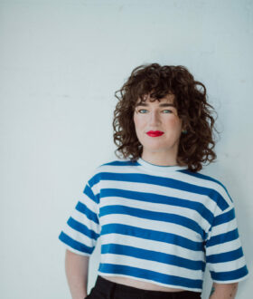 A woman with brown curly hair and bright red lipstick, wearing a striped blue and white shirt, leans against a white background