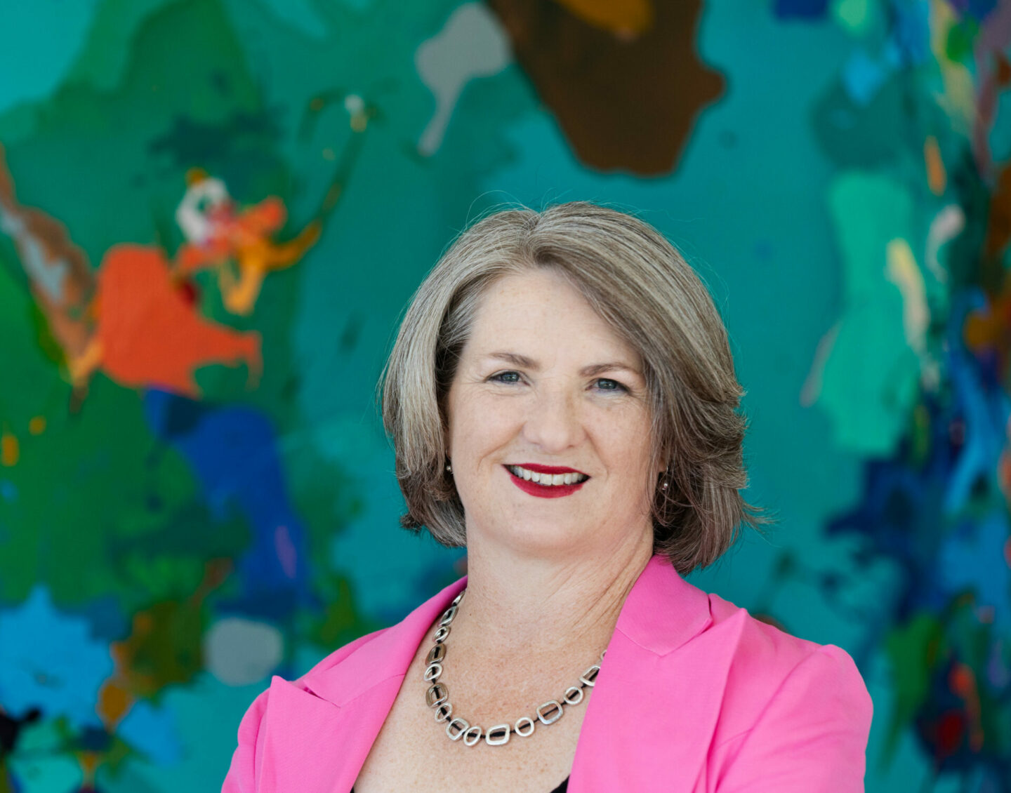 A short-haired white woman in a pink blazer smiles in front of a colourful background