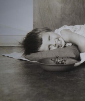 A black and white photograph of a smiling child lying on a pillow on the floor behind a bowl