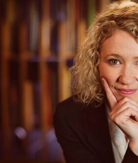 A curly-haired blonde woman leans on her hand and smiles, looking directly at the camera