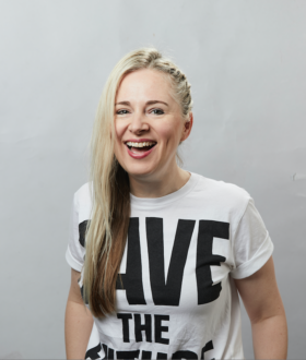 A laughing woman with long blonde hair falling over one shoulder, wearing a white t-shirt with black lettering that reads Save The Future
