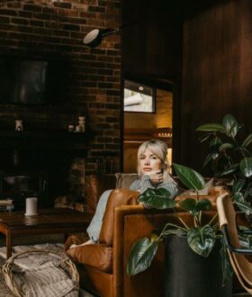 A photograph of a blonde woman sitting on a sofa in a lounge room, her body obscured by the side of the sofa and a pot plant