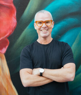 A man with short gray hair and red-rimmed glasses stands arms-folded and smiling in front of a colourful artwork.