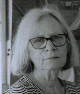 A black and white headshot of a woman with chin-length gray hair and glasses, looking at the camera without smiling