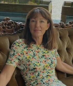 A middle-aged woman with a floral dress leans against a brown couch. She has mid-length brown hair and a fringe