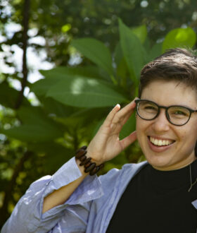 A person with short brown hair smiles and touches both hands to their dark-rimmed glasses, against a backdrop of green plants