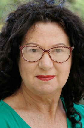 A close-up of a middle aged woman with dark curly hair and red-rimmed glasses
