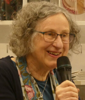A grey-haired woman with glasses and a floral scarf leans forward an speaks into a handheld microphone