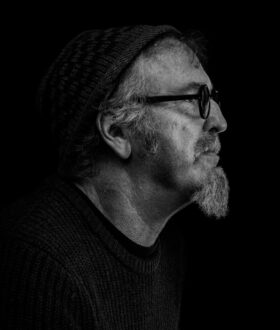 A black and white photo of a side-on view of a man with a beanie, dark-rimed glasses and a goatee