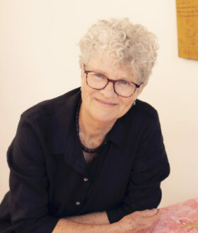 A woman with short curly gray hair and brown-rimmed glasses leans forward over a table, resting on her folded arms