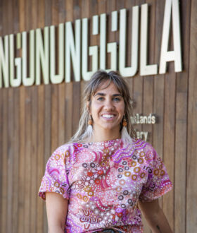 A brown-skinned woman with straight grey-brown hair smiles, leaning against a barrier, wearing a pink top patterned with Aboriginal art