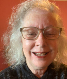 A close-up photograph of an older woman with shoulder-length gray hair and brown glasses, laughing as she leans towards the camera