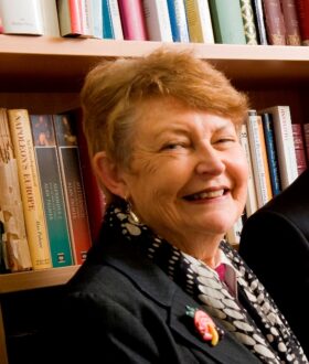 An older woman with short red hair turns side on to the camera and smiles, in front of a bookshelf