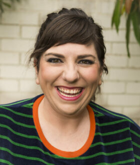 A photograph of a woman with brown hair worn up with a fringe and amber eyes, wearing a green and blue striped shirt and smiling widely