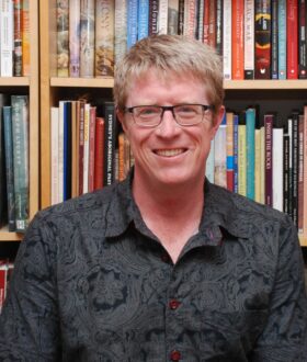 A blond man with dark-rimmed glasses and a black shirt smiles in front of a bookshelf