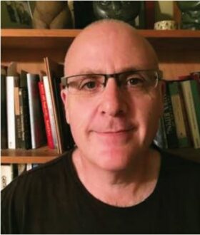 A bald man with thin-rimmed glasses and a black tshirt smiles closed-lipped in front of a bookshelf