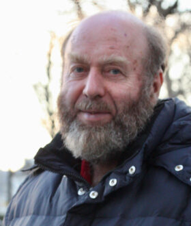 A photograph of a bald man with a bushy gray beard standing tilted away from the camera in a thick jacket