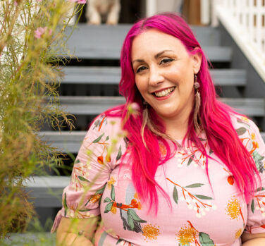 A woman with long bright-pink hair and a pale pink floral dress sits on some steps and smiles