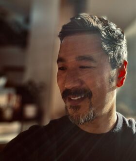 An Asian man with a goatee stands at an angle, smiling, as light shines across his face