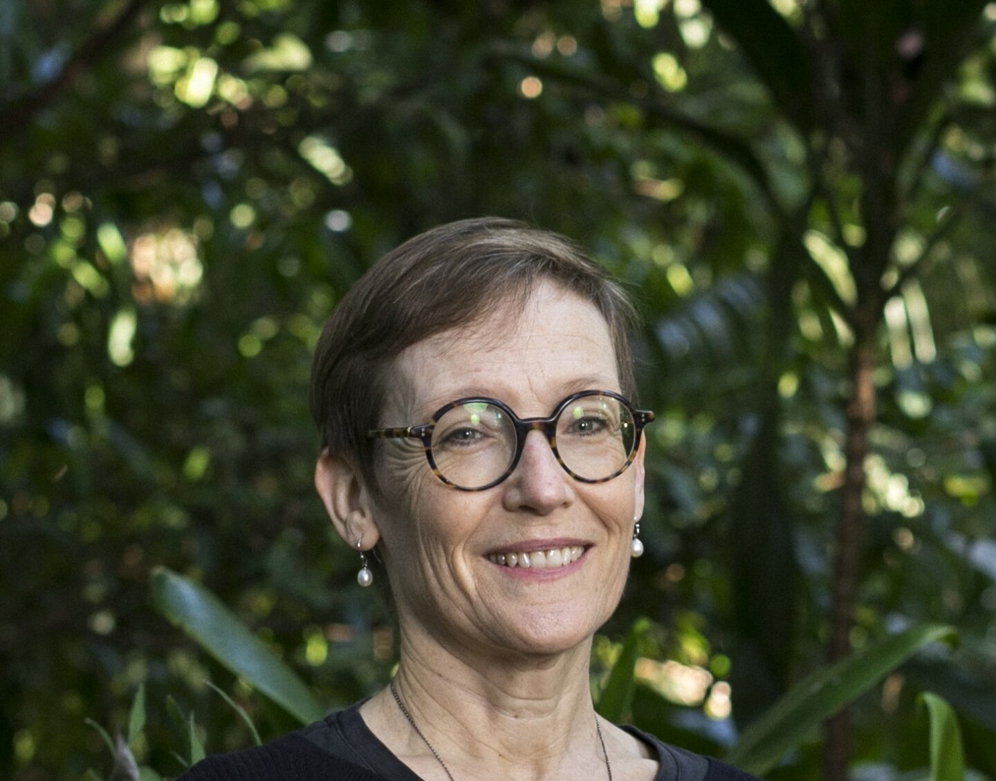 A middle-aged woman with short brown hair and brown, round glasses smiles in front of a tree