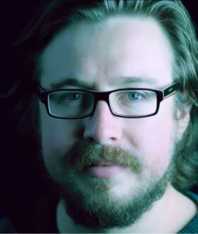 A close-up photograph of a young man with shoulder-length wavy hair, a medium-length brown beard, and rectangular black-framed glasses
