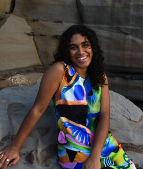 A dark-skinned woman with long curly black hair sits on a rock, smiling widely in a colourful dress