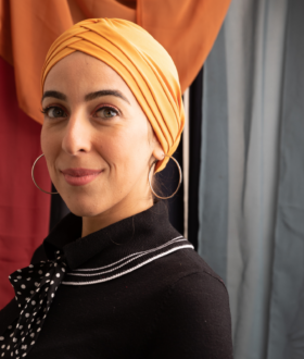 A woman with large hoop earrings and a yellow headscarf stands at an angle to the camera, smiling with closed lips
