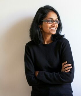 A dark-skinned woman with shoulder-length black hair and glasses stands arms folded, looking to one side and smiling widely