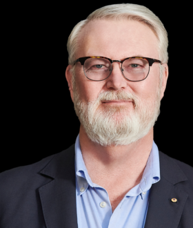 A man with white hair and full, trimmed beard, wearing dark-rimmed glasses and a dark gray blazer