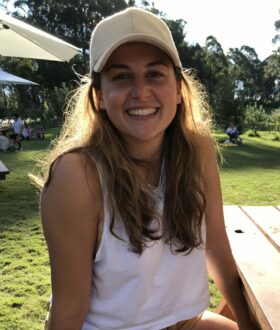 A young woman with long brown hair and a white cap smiles at the camera, sitting at a picnic table