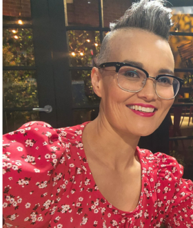 A woman with a short gray mohawk and dark-rimmed glasses leans into the frame, smiling