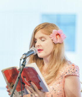 A blonde woman with a pink flower in her hair reads from a book at a microphone
