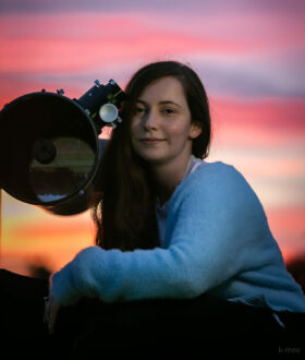 A young woman with long brown hair sits beside a telescope, the sky behind her is purple and pink