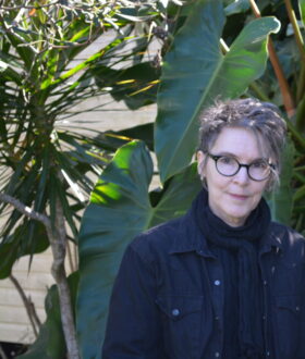 A white woman with round glasses and hair worn loosely up, standing in front of a plant with large leaves