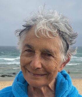 An older woman with short grey hair and a fleecy blue jacket stands smiling on a beach