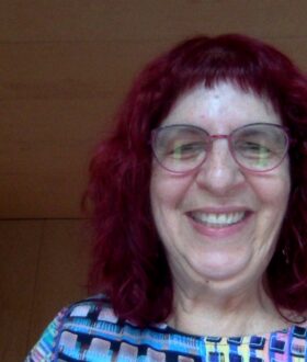 A white woman with curly dark red hair and glasses smiles widely