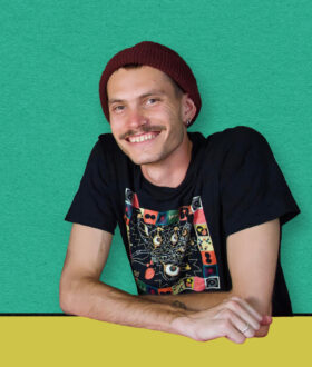 A young white man with a moustache, nose and ear piercings and red beanie leans over a yellow desk and smiles